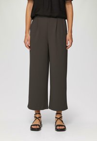 Donkerbruine wijde broek met plooien, gemaakt van een zachte stof. Draagt met zwarte sandaaltjes met bandjes, die de enkels laten zien.