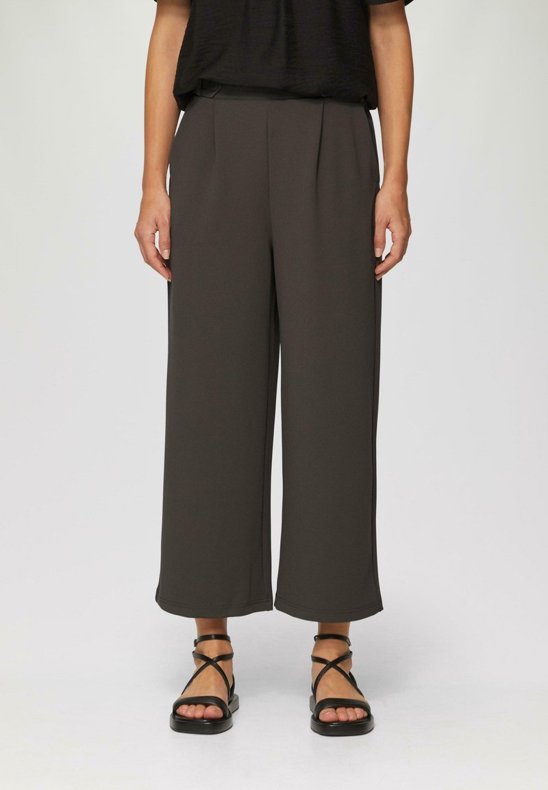Donkerbruine wijde broek met plooien, gemaakt van een zachte stof. Draagt met zwarte sandaaltjes met bandjes, die de enkels laten zien.