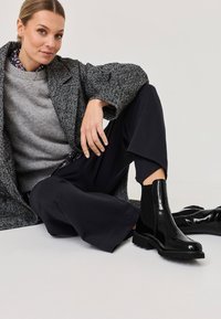 Černé lakované kotníkové boty s elastickými bočními panely a silnými gumovými podrážkami, v kombinaci se šedým svetr a texturovaným šedým kabátem.
