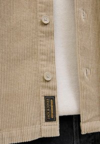Beige corduroy blazer met twee knopen, voorzien van een merketiket. Daaronder zijn een lichte gebreide top en donkere denim zichtbaar.