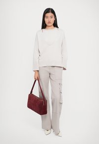 Kvinde iført en lys beige sweater og cargobukser, som holder en burgunder håndtaske i venstre hånd, stående foran en helt hvid baggrund.