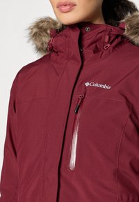 Bordeauxfarbene, wasserdichte Jacke mit Kapuze aus Kunstpelz, mit Reißverschluss auf der Brust und Columbia-Logo auf der Brust. Glatte Textur und warmes Design.
