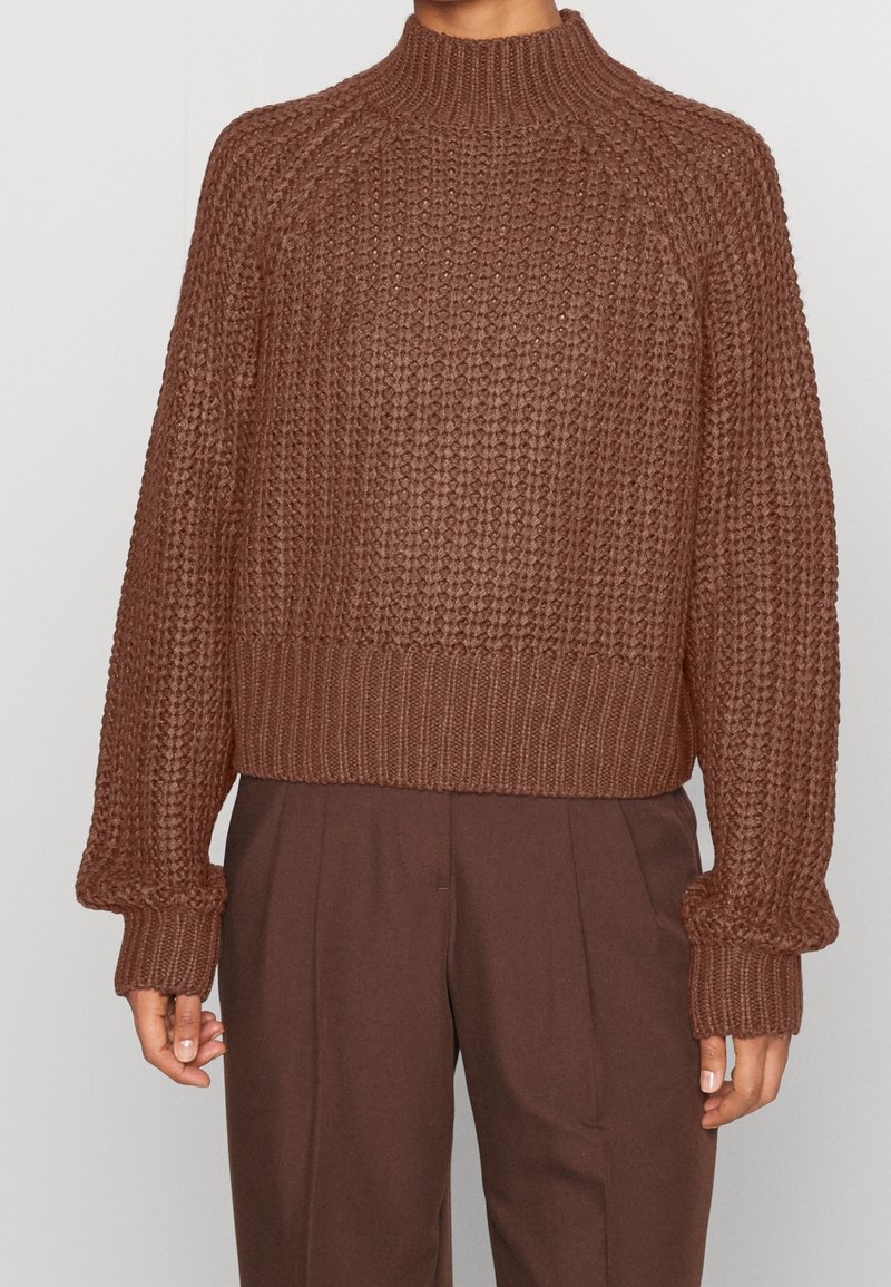 Brown knitted sweater with a high collar, ribbed hem, and wide sleeves featuring a textured pattern. Paired with brown trousers.