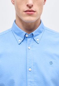 Άνδρας που φοράει γαλάζιο πουκάμισο με κουμπιά και μικρό κεντημένο λογότυπο "S" στο στήθος, απεικονίζεται από το πηγούνι μέχρι το μέσο του στήθους.