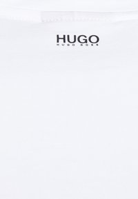 Fehér pamut póló, kerek nyakkivágással. A "HUGO" márka logója fekete színben szerepel a "HUGO BOSS" felirat felett elöl.