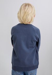 Niño rubio de espaldas, vestido con una sudadera azul marino y jeans de un azul claro, contra un fondo claro y liso.
