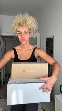 Δύο στοίβες κουτιά που κρατούνται, το ένα καφέ από χαρτόνι με το λογότυπο της Zalando και το άλλο άσπρο. Ο χώρος έχει ανοιχτόχρωμους τοίχους και έναν καθρέφτη στο background.