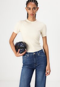 Kremfarget, ribbestrikket topp med korte ermer kombinert med høyt liv, blå denimjeans. Modellen holder en sammenfoldet mørkeblå veske. Enkelt, rent design.
