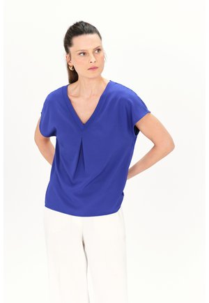 Blusa blu royal a maniche corte con scollo a V e dettaglio plissettato nella parte anteriore, abbinata a pantaloni bianchi a gamba larga su uno sfondo neutro.