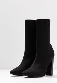 Černé pletené kotníkové boty s špičatou špičkou, žebrovaným lemem a vysokým blokovým podpatkem. Materiál má těsnou, hladkou texturu. Žádné ozdoby.