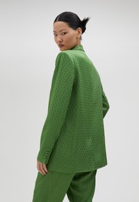 Traje verde con un patrón, que incluye una americana con cuello y puños abotonados, combinado con pantalones a juego. Textura suave, ajuste a medida.