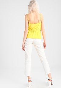 Top sin mangas amarillo con diseño de espalda fruncida, combinado con jeans cortos blancos. La prenda cuenta con tirantes finos y un corte relajado.