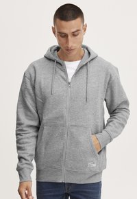 Man wearing light grey zip-up hoodie with front pockets and drawstrings, looking down, with right hand in pocket and left arm relaxed.