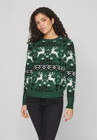 Pull en maille verte avec des rennes blancs, des flocons de neige et le texte "JOLI NOËL !". Encolure ronde et poignets et ourlet côtelés.