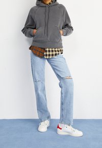 Felpa grigia con cappuccio, indossata sopra una camicia a scacchi marrone e nera. Jeans azzurri sbiaditi e scarpe da ginnastica bianche con accenti rossi.
