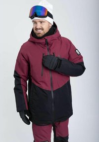 Marsala- og sort skijakke med hætte, udstyret med lynlåslommer og logoer. Model iført goggles og handsker står foran en neutral baggrund.