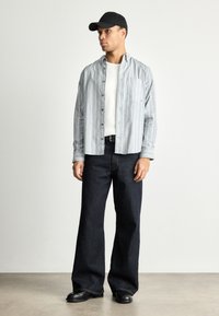 Pelēkas svītrains krekliņš ar pogām virs balta T-krekla, apvienots ar tumši zilajiem platmalu džinsiem un melnām kurpēm. Stilizēts ar melnu cepuri.