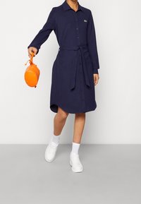 Marinblå långärmad skjortklänning med knappar, knytning i midjan och Lacoste-logga. Modellen håller en liten orange väska, vilket matchas med vita sneakers.