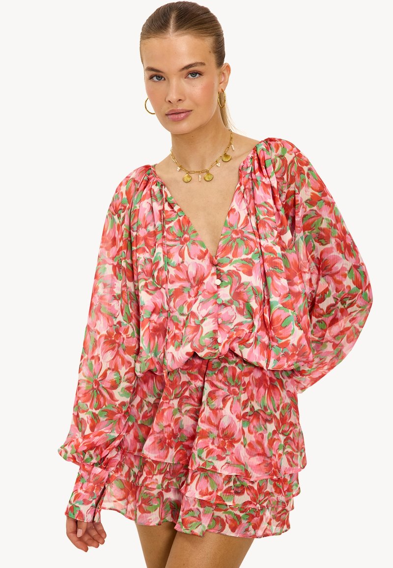 Vrouw in een roze bloeiende blouse met lange mouwen en pofmouwen, gecombineerd met een bijpassende mini-rok met gestapelde ruches, accessorized met een gouden schelpenhanger en oorringen.