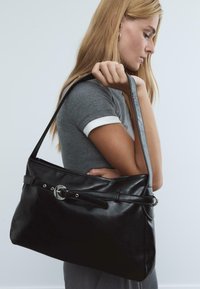 Junge Frau mit langen blonden Haaren, die ein graues Shirt trägt, hält eine große schwarze Ledertasche mit einer Schnallenverzierung.