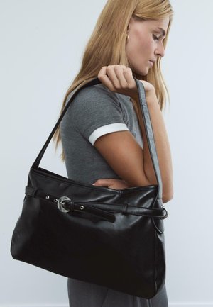 Jeune femme aux longs cheveux blonds portant une chemise grise tient un grand sac à épaule en cuir noir avec un détail de boucle.