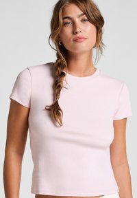 Jonge vrouw met gevlochten haar, gekleed in een lichtroze, getailleerde, kortdangende top tegen een effen achtergrond.