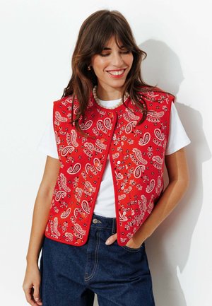 Femme souriante les yeux fermés, portant un gilet rouge à motif cachemire sur un t-shirt blanc et un jean bleu foncé, main dans la poche.