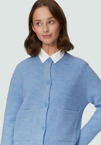 Helles blaues Cardigan aus strukturiertem Strickstoff. Verfügt über eine Knopföffnung, zwei Fronttaschen und ein weißes Hemd mit Kragen darunter.