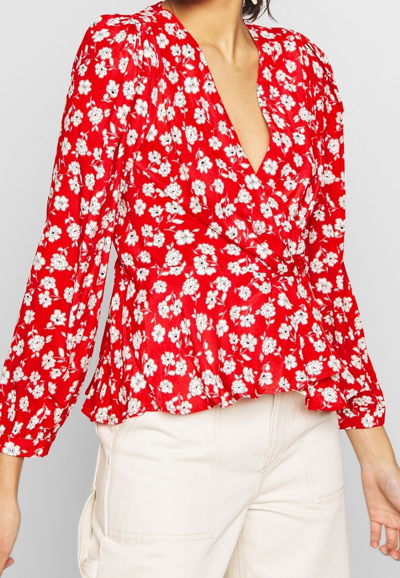 Femme portant un chemisier cache-cœur rouge à fleurs blanches avec manches longues et un pantalon couleur crème, visible des épaules jusqu’aux hanches.