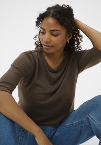 Brązowa bluzka z krótkim rękawem, wykonana z miękkiej tkaniny o luźnym kroju. Noszona z niebieskimi dżinsami. Minimalistyczny design, bez widocznych wzorów.