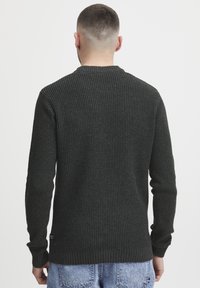Mørk grønn strikkegenser med ribbestrikket hals og mansjetter, med et strukturert mønster og et lite logo-patch på den nedre siden.