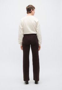 Barna kordbársony trapéznadrág hátsó zsebekkel, világos bézs pulóverrel párosítva. Modell hátulnézetben, semleges háttér előtt.