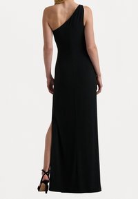 Svart, figurnära klänning med en axel och en sidslits. Slät, elastisk tyg; elegant silhuett som framhäver figuren. Svarta högklackade skor.