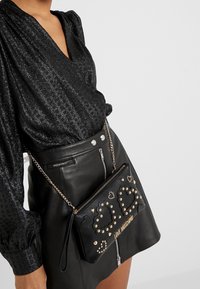 Černá kožená crossbody taška se zlatými hroty, srdíčky a logem značky. Obsahuje řetízek na rameno a možnost rukojeti.