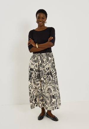 Mujer sonriente con el pelo corto de pie con los brazos cruzados, lleva una camiseta negra, falda floral en blanco y negro, pendientes de aro dorados, pulsera y zapatos planos con tachuelas.