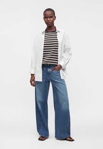 Personne debout portant une chemise blanche boutonnée sur un haut à rayures, un jean bleu large et des sandales, sur un fond uni et clair.