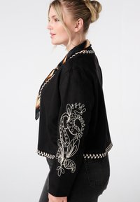 Blonde vrouw in profiel met een zwart jack met witte bloemembroideries op de mouwen en de zoom, zwarte jeans, grote hoopoorbellen en een gedessineerde sjaal.