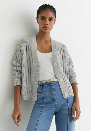 Femme aux cheveux tressés porte un cardigan en tricot gris clair par-dessus un débardeur blanc et un jean bleu taille haute avec une ceinture beige.