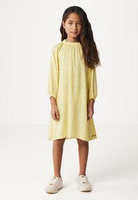 Gelbes Kleid mit Blumenmuster, langen Ärmeln und gerafftem Ausschnitt. Getragen mit weißen Turnschuhen, die eine strukturierte Sohle und Schnürsenkel haben.