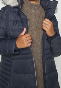 Marineblaue Steppjacke mit Kunstfellkapuze, versehen mit einem Reißverschluss und gerippten Bündchen, getragen über einem braunen Rollkragenpullover. Zwei Ringe an den Händen.