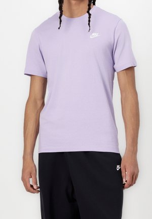 Person iført en lyse lilla Nike T-shirt og sorte Nike bukser står mod en ensfarvet hvid baggrund.