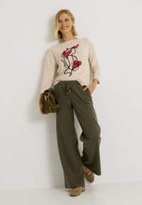 Chemise beige à manches longues avec un design graphique rouge et noir, associée à un pantalon ample vert olive et des chaussons beiges avec des motifs découpés.