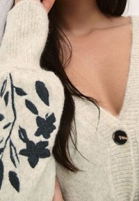 Ljusgrå kofta med djup V-ringning, med mörk blommig broderi på vänster ärm och en rund svart knappstängning.