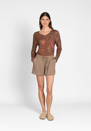 Woman standing with hands in pockets wearing a long-sleeve floral top, beige striped shorts, and beige slippers on a plain background.