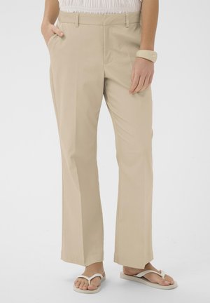 Beige gerade geschnittene Hose, kombiniert mit weißen Flip-Flops, Hand in der Tasche und beigem Armband am Handgelenk, von der Taille bis zu den Füßen gezeigt.