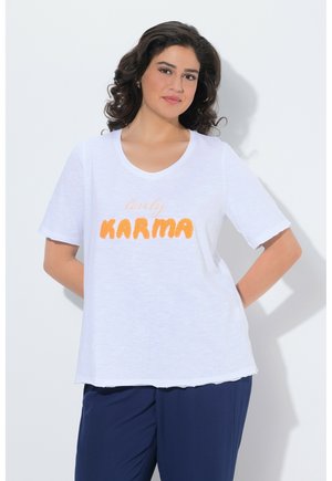 Kvinde med krøllet mørkt hår iført hvid T-shirt med orange "lovely KARMA" tekst og marineblå bukser, stående med hænderne bag ryggen.