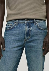 Taillenteil einer Person in hellblauen Jeans und einem beigen, strukturierten, ärmellosen Oberteil, die Hände entspannt an den Seiten.