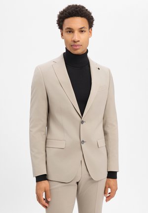 Junger Mann, der einen beigen Anzug mit schwarzem Rollkragenpullover trägt, vor einem schlichten weißen Hintergrund stehend und nach vorne blickend.