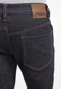 Jeans de mezclilla oscura con una etiqueta de cuero marrón visible en la cintura. Presenta costuras clásicas y bolsillos traseros estándar.