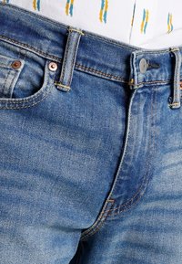 Siniset farkut klassisella designilla, joissa on vetoketjusuljin, kupariset niitit ja näkyvät saumat. Kangas on hieman haalistuneen tekstuurinen.